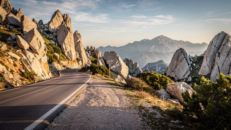 Route de montagne sinueuse près de Bavella, en Corse-du-Sud, avec un panneau discret et un paysage minéral rocheux, éclairage doré de fin de journée.