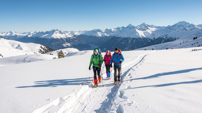 Deux femmes et un homme en raquettes avancent sur un sentier enneigé du plateau de Solaison, entouré d’alpages blancs sous la lumière hivernale.